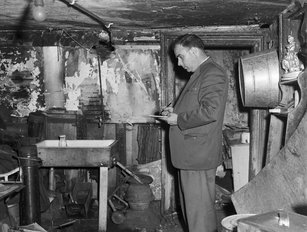 Black and white image of inspector writing in notepad in basement surrounded by debris.