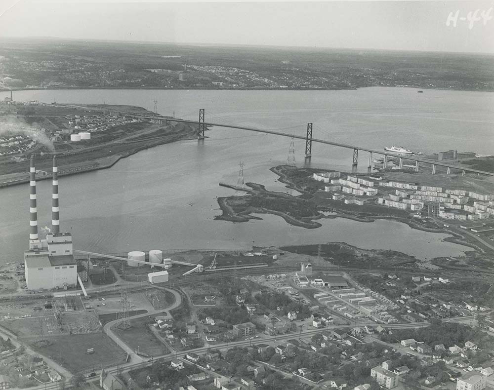 Black and white oblique aerial photograph looking northwest from Windmill Road towards the narrows and MacKay Bridge, showing 2 stacks at the Tuft's Cove generating Station, Shannon Park the Bedford Institute of Oceanography and across the Harbour to a demolished Africville and the Rockhead city prison lands. 