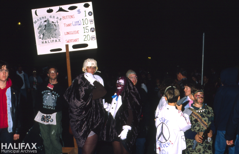 Colour photo of costumed partiers including vendor selling Mardi Gras Halifax Tshirts, buttons, etc.