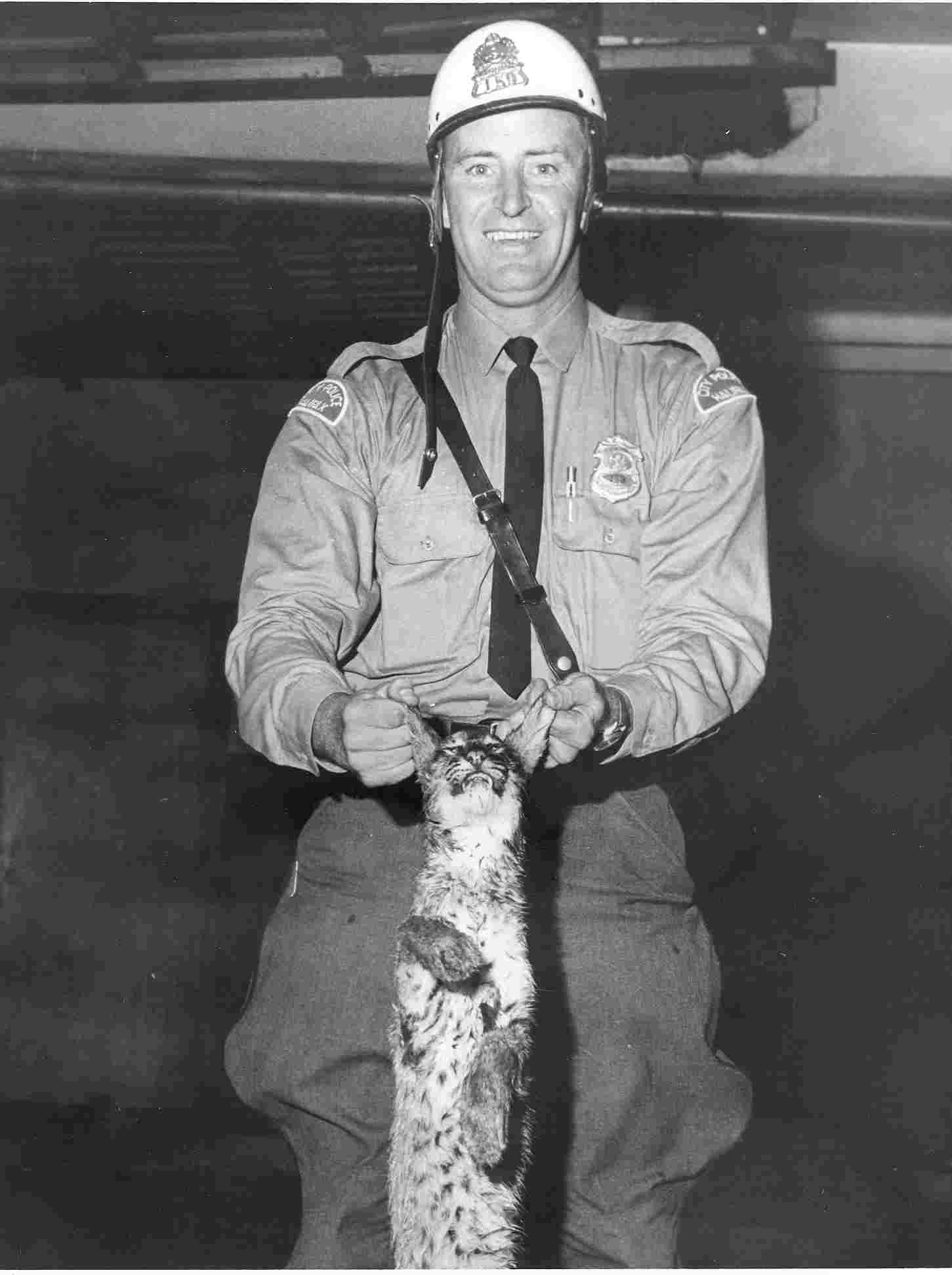 Black and white photo of police officer smiling holding wildcat by the ears