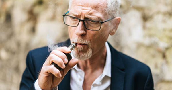 Photgraph of a mature man with gray hair and black glasses wearing a blue blazer jacket and white shirt and smoking an e-cigerette.  Smoke is puffing from it.