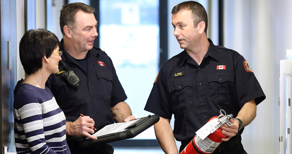 Close-up image of two Halifax Regional Fire & Emergency Station Officers conducting a Fire Safety Maintenance Inspection of a building while having a friendly conversation with a female property representative. One Officer is holding a portable fire extinguisher.