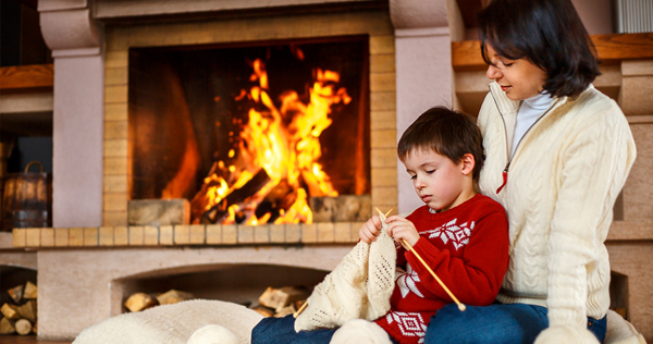 Image of a mother and young son sitting on a large cusion in front of a fire in a fireplace while the Mom is teaching her son to knit.