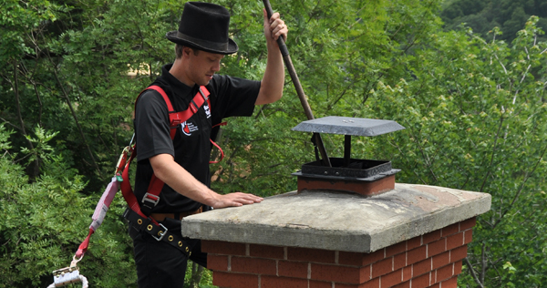 Close up image of a man wearing an old style chimney sweep top hat and a modern safety harness up on the roof on a building cleaning a chimney flue