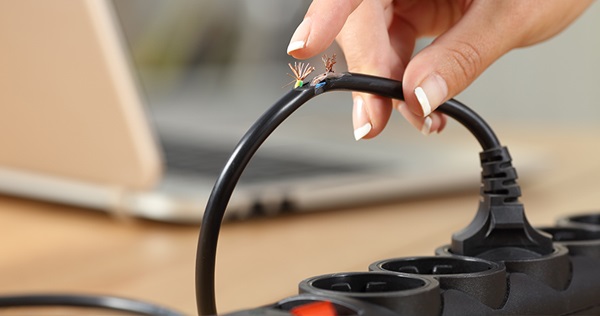 Close up image of a woman's hand holding an electrical cord plugged into a power bar and her finger is just about to touch exposed charged wires on the frayed cord.