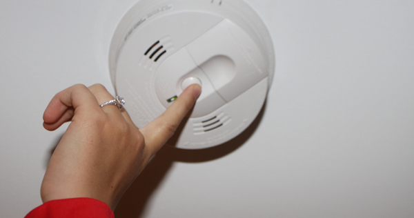 Close up image of a womans finger pushing the test button on a ceiling mounted smoke alarm