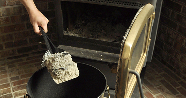 Close up image showing a woodstove with the door open and a persons hand holding a small metal shovel is removing the ashes from the woodstove and placing them in a black metal ash bucket.
