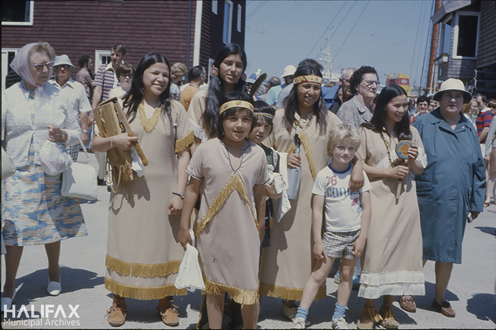 Image of a group of Mi'kmaq in traditional dress posing with a family during Natal Day celebrations on the Halifax waterfront