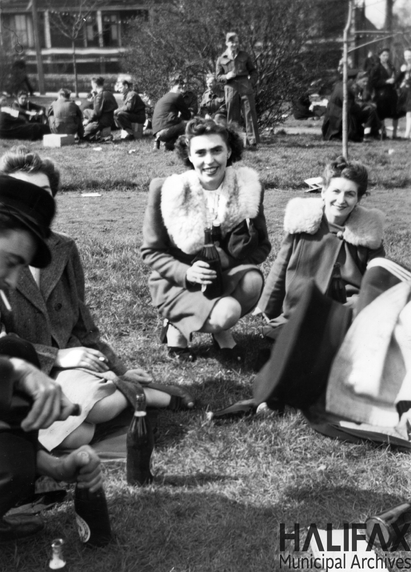Black and white photograph of 3 young women and a soldier drinking and smoking sitting on the grass .