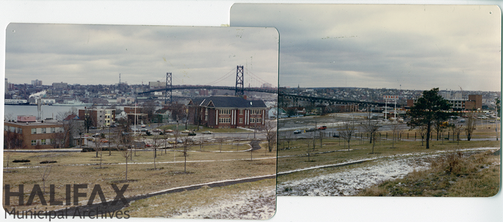 Colour photograph from a hilltop showing parkland, a parking lot, a large brick building, and a bridge in the background, and a smattering of snow lying gently upon the grass in the foreground.