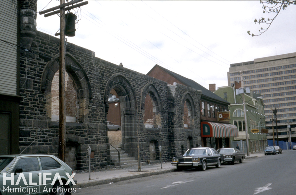 Colour photograph showing street with parked cars, buildings, and the first storey facade of a stone building