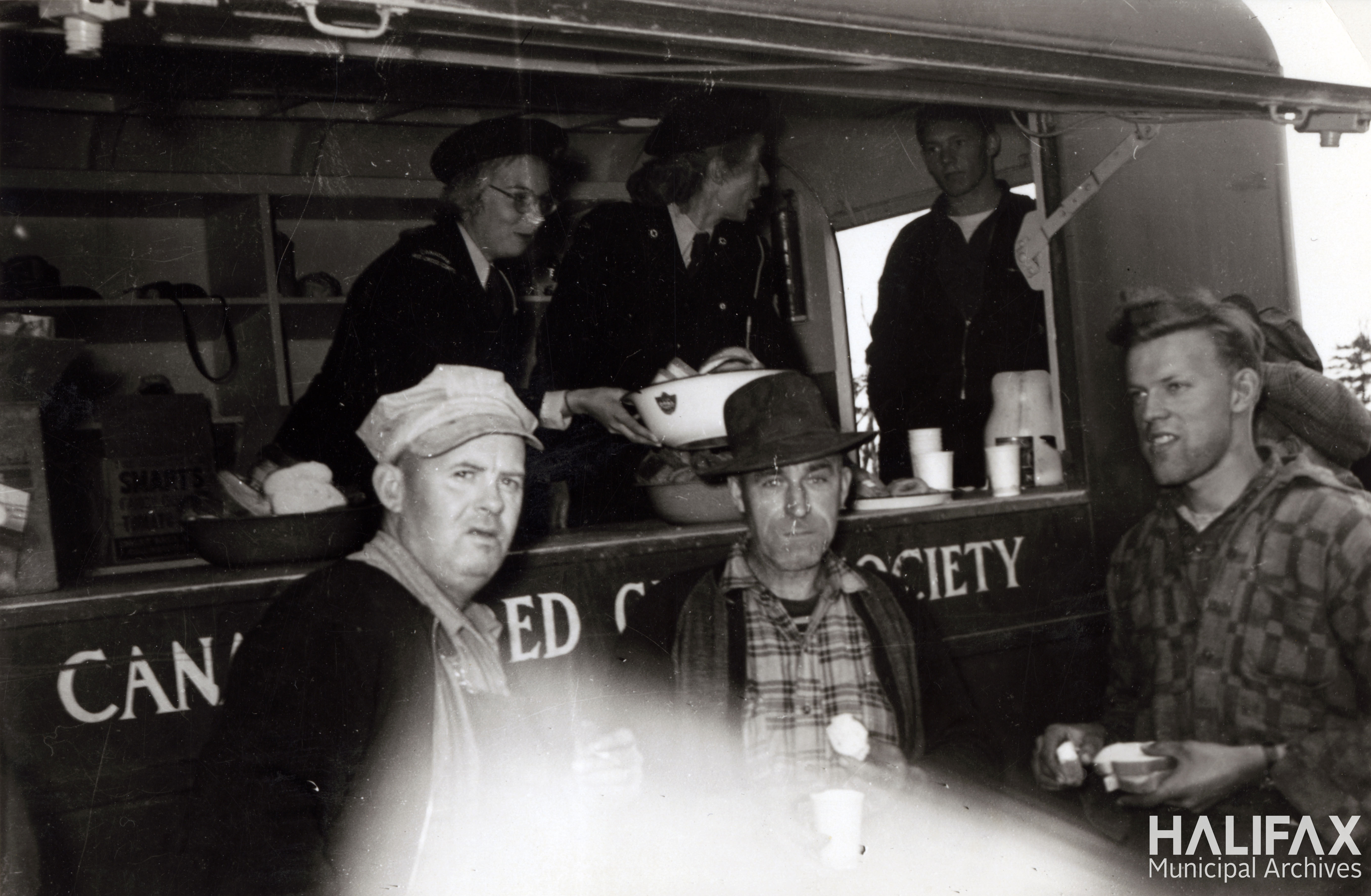 Black and white image of uniformed women working in a mobile kitchen serving 2 firemen waiting outside of van.