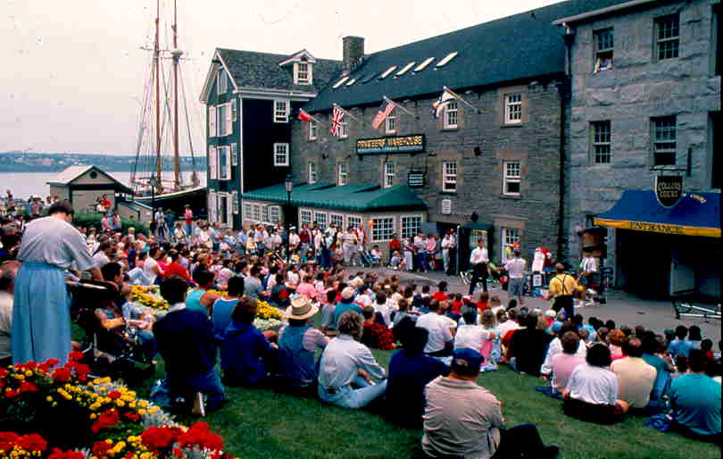Colour photo of buskers entertaining crowds with schooner in background.