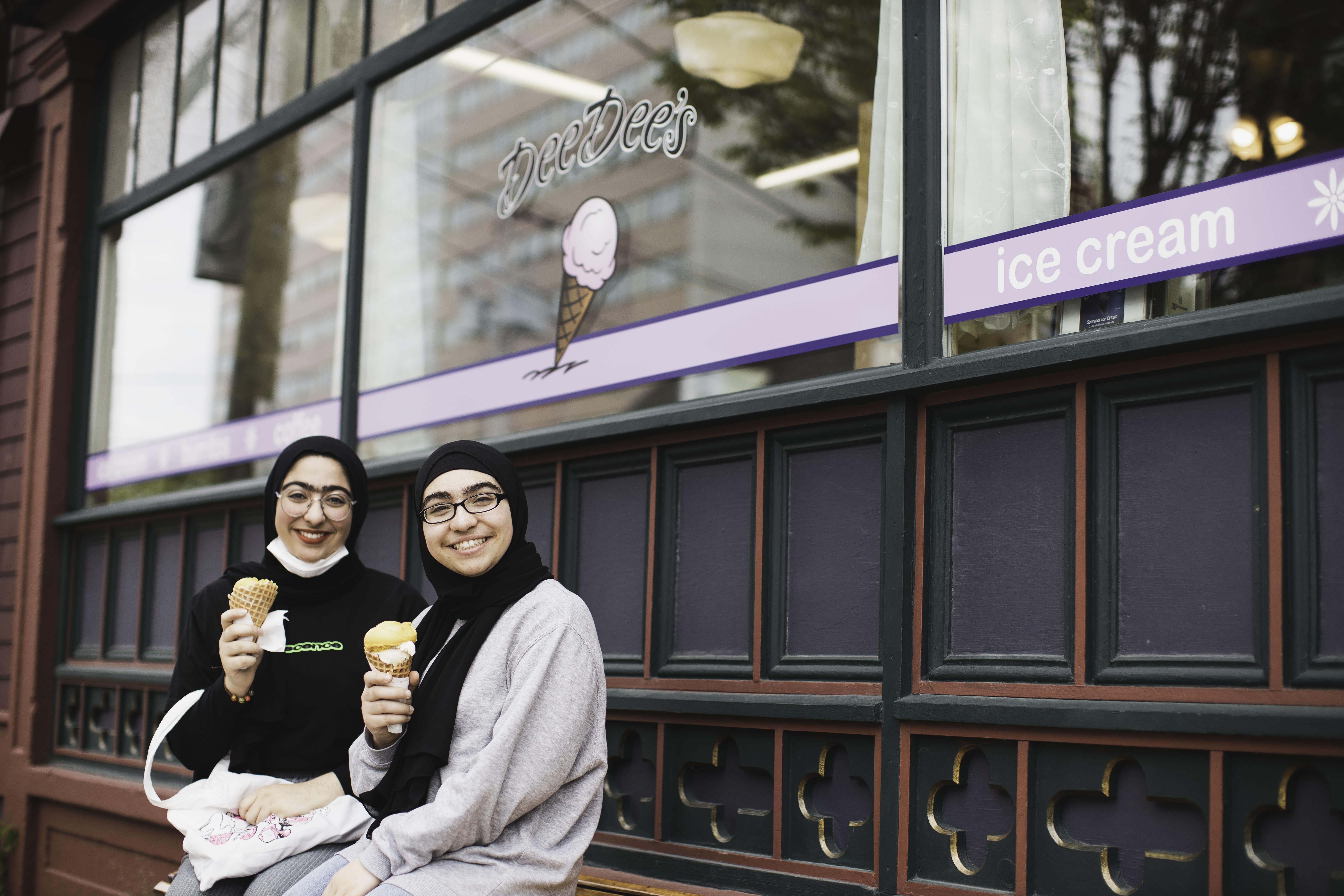 Pictured: two young women sitting outside DeeDee's ice cream shop in Halifax eating ice cream and smiling