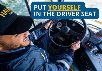 A male bus driver sits behind the wheel of the bus. He's wearing a baseball cap that says Halifax Transit and his hands are on the wheel. 