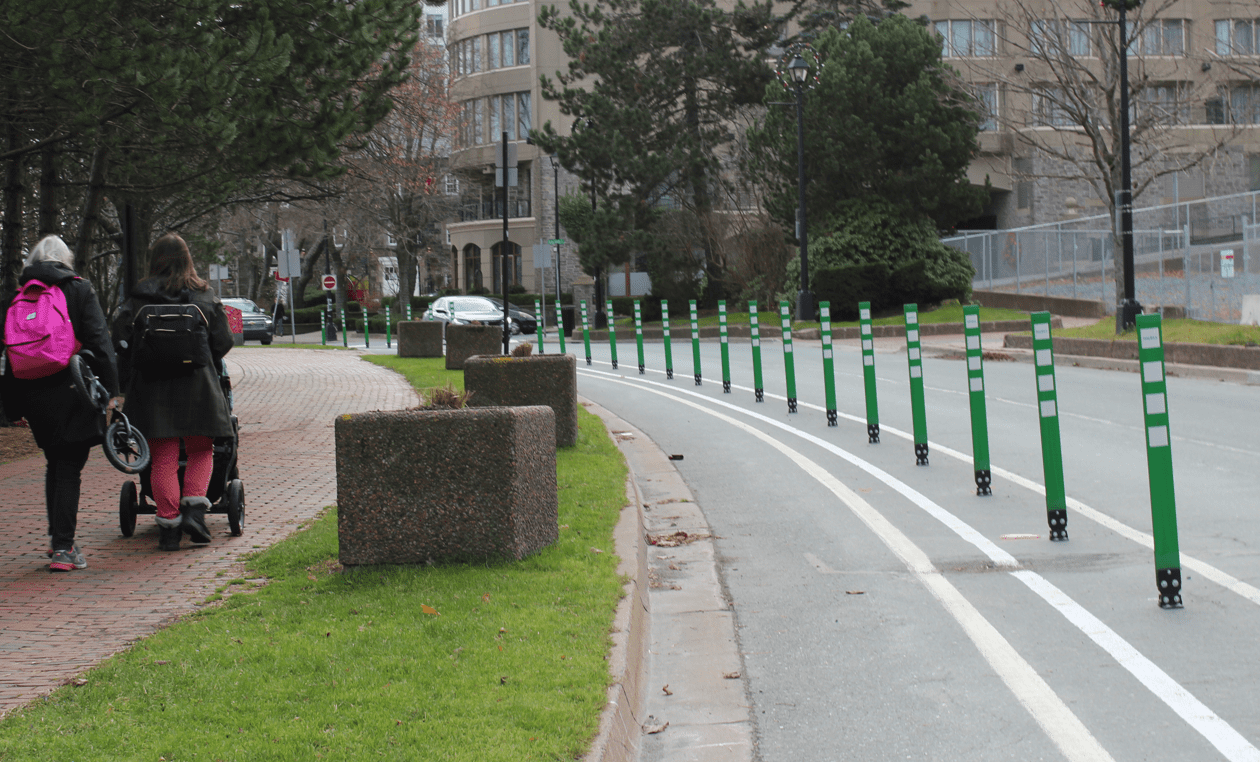 A photo of the Lower Water Street Bollard-Protected Bikeway. On the left of the photo is a sidewalk with two pedestrians walking away from the camera. On the right is Lower Water Street with the bollard-protected bikeway on the left side of the street and a vehicle lane on the right.