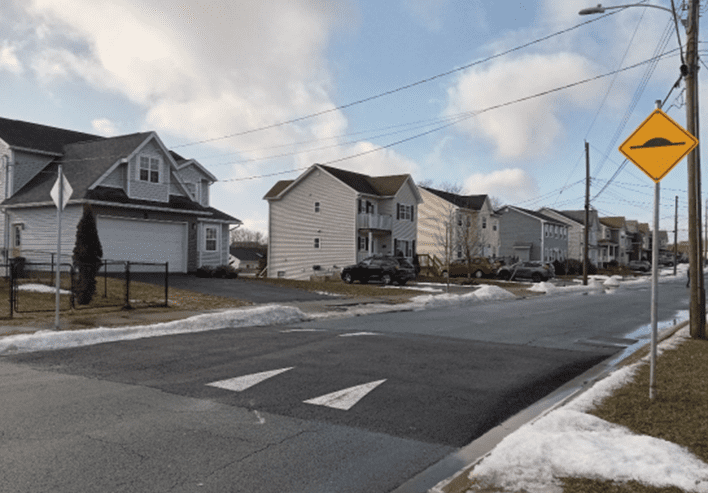 Photo of a residential street lined with single family houses. The street features a speed table, designed similar to a longer speed bump, with white arrows painted on it and a sign cautioning drivers.