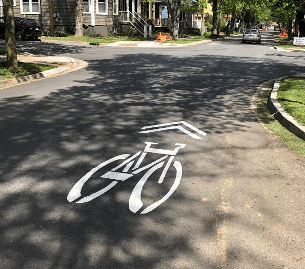 A close-up photo of a street with a white bicycle and two sharrows painted on it to advise cyclists of a street where they can ride single file with vehicle traffic without a designated bicycle lane. 