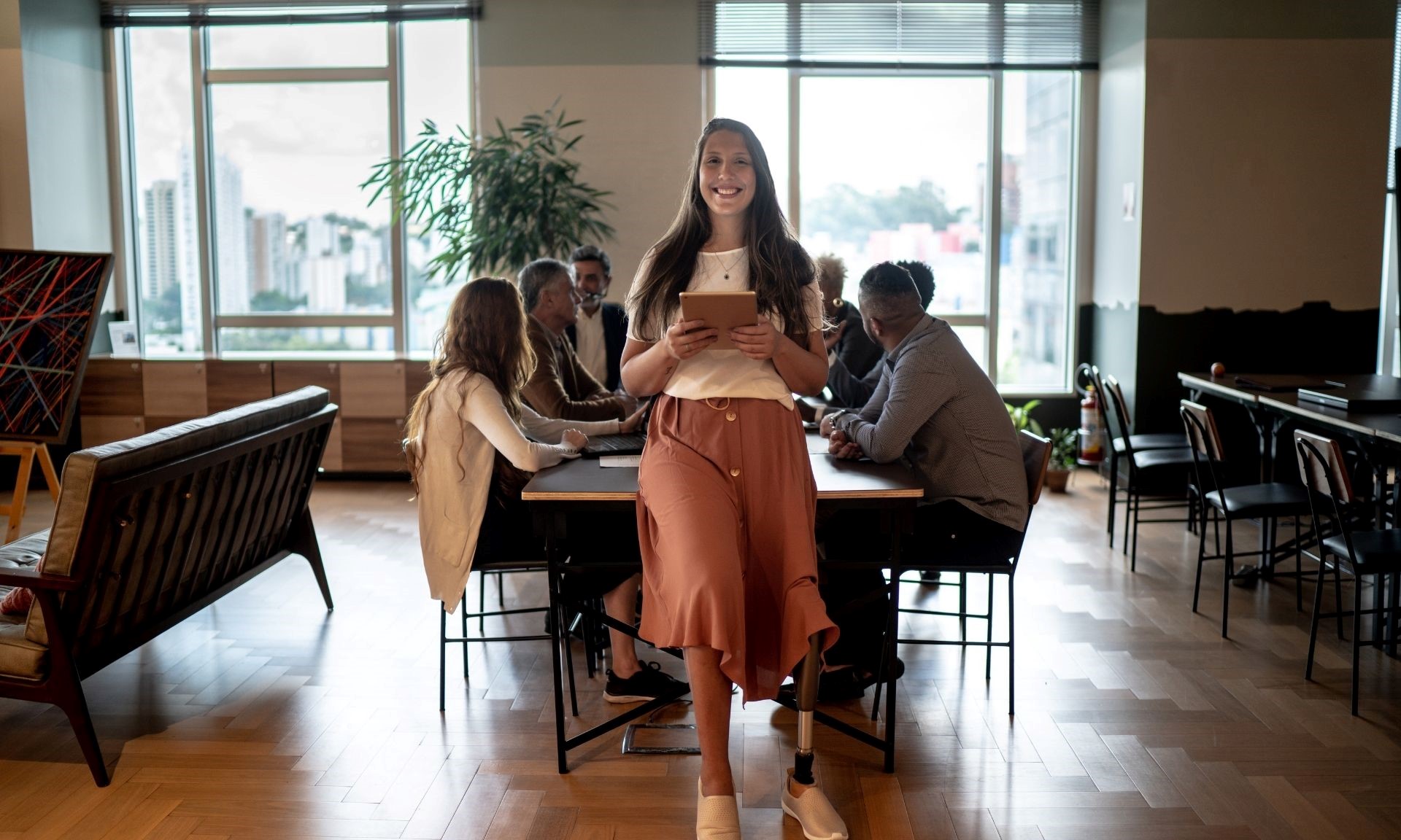 A young woman sits at the end of a board room table during a meeting. We can see that she has a prosthetic leg.