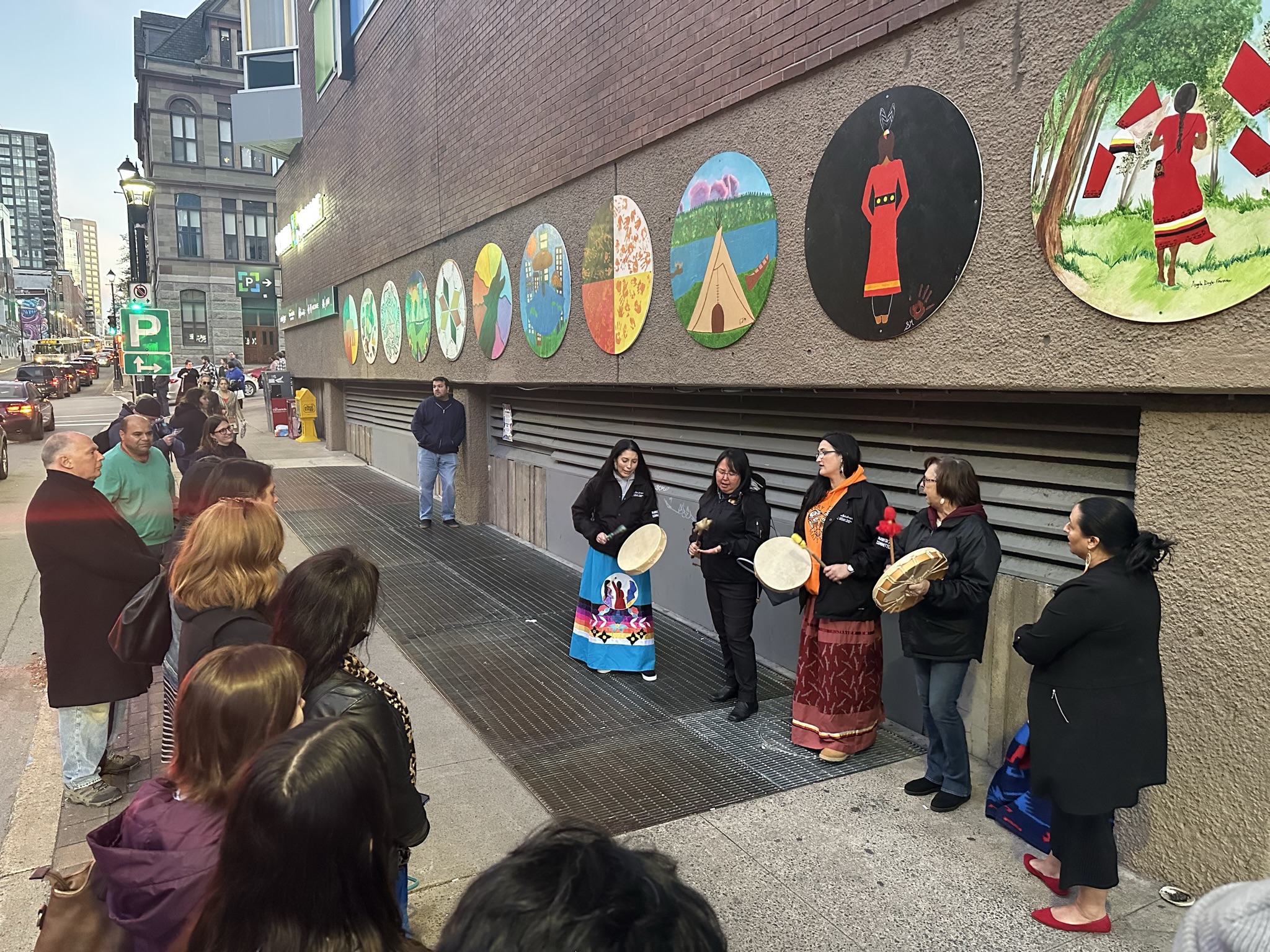 A crowd is gathered around the Our Stories murals on Barrington Street. People perform Indigenous traditions in front of the murals.