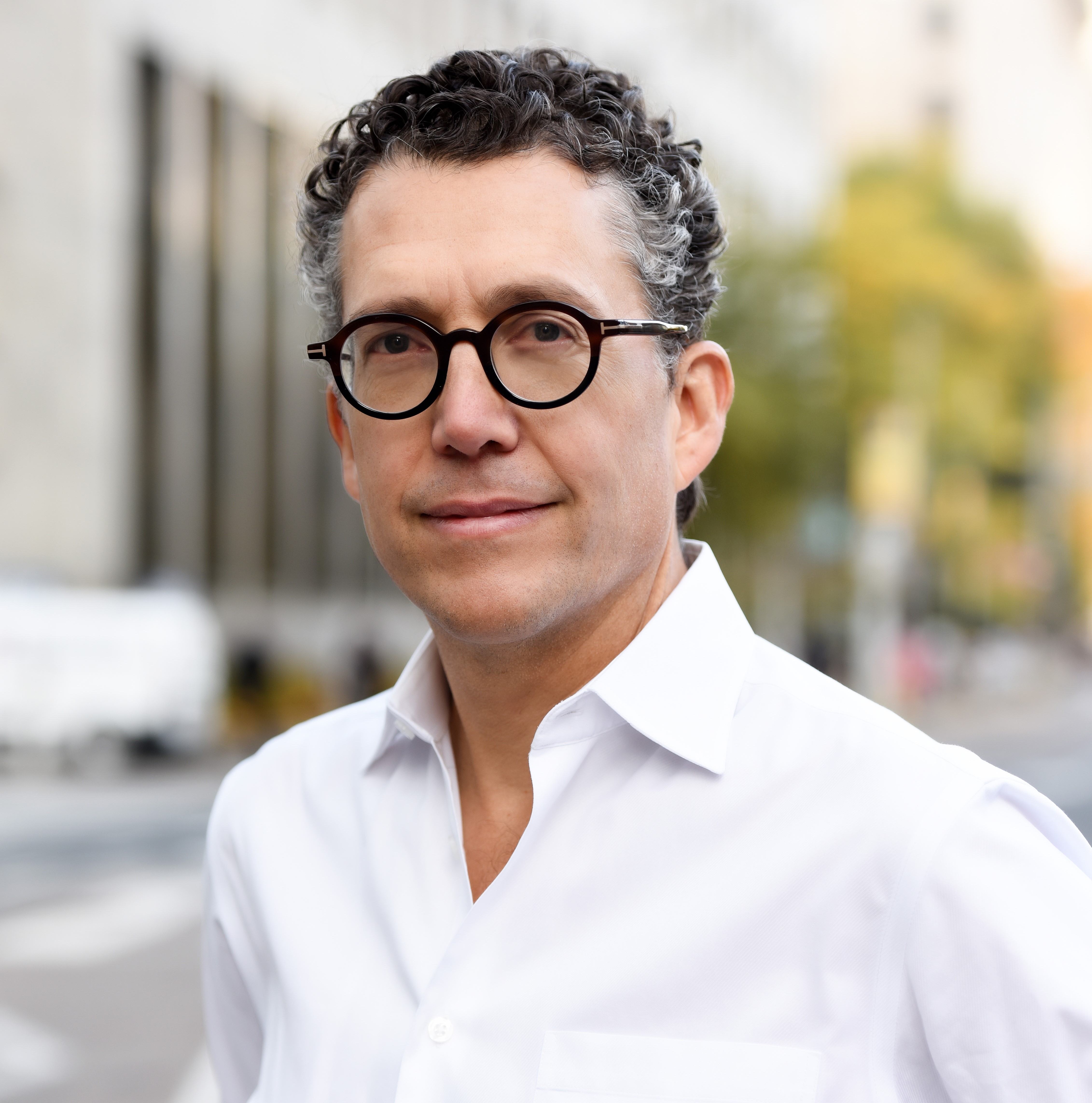 Headshot of Antonio Gomez-Palacio, architect and urban planner. Antonio wears a white shirt and black glasses. He is standing outside, with a streetscape in the background.