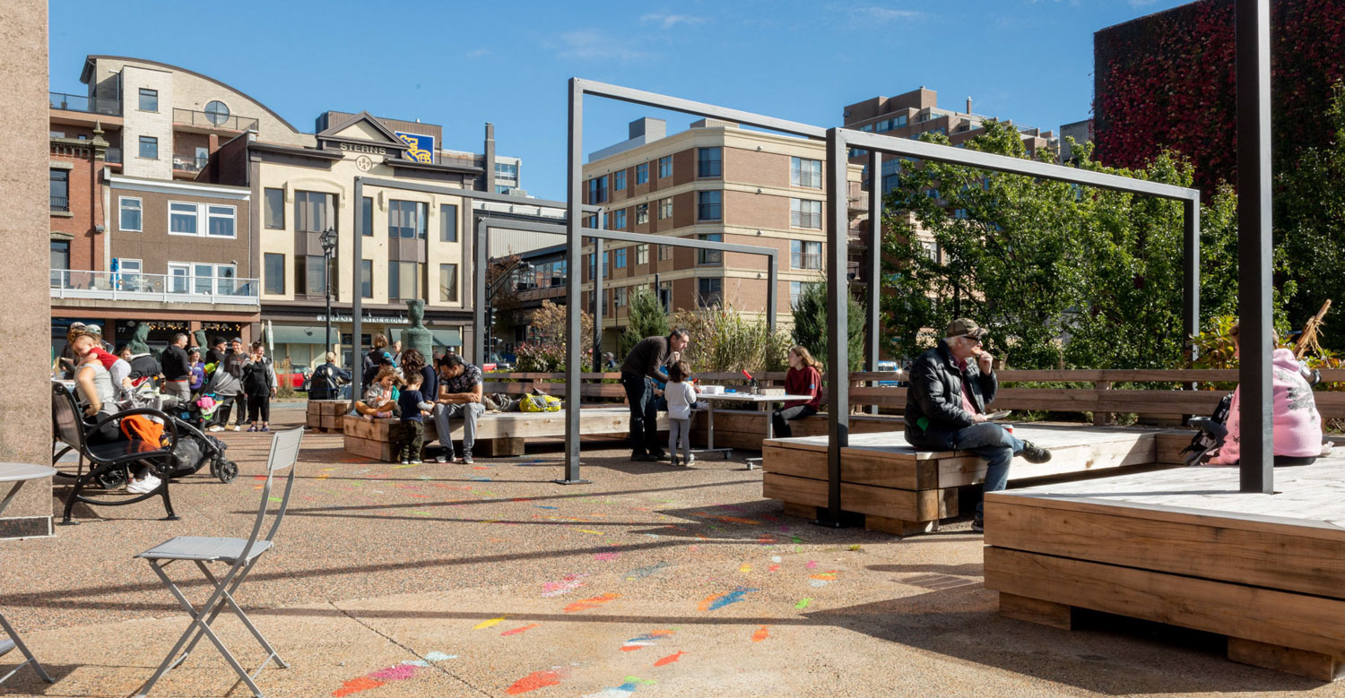 Alderney Plaza on a sunny summer day. There are square wooden benches, fold-out patio chairs and tables, and the ground is painted with a colourful design.