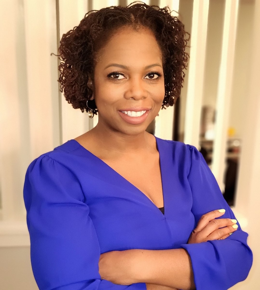 Headshot of Camille Mitchell, architect. Camille is standing with her arms crossed, is wearing a blue shirt and is posing against a beige background.