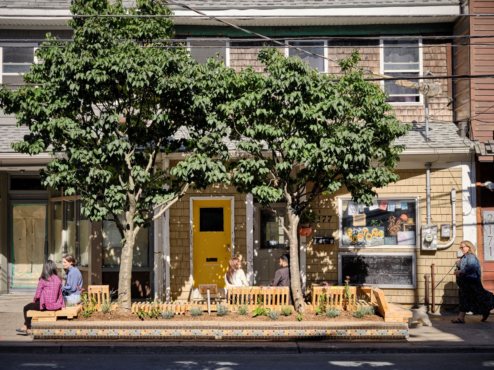 Wooden bench located on Gottingen St in front of a local business. People are sitting on it to the left side and there are trees in the background. It is a sunny day.