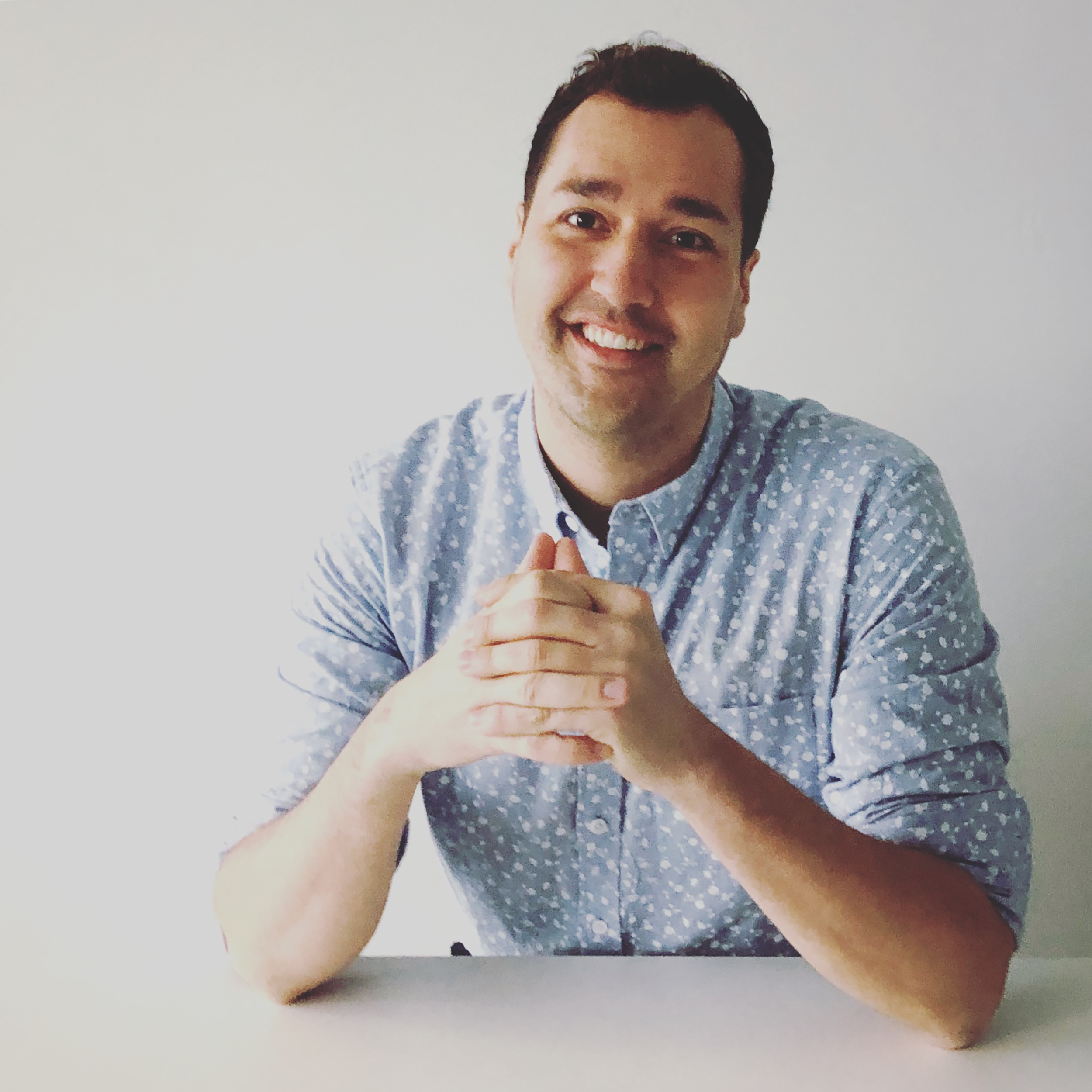 Headshot of Matthew A.J. Brown, landscape architect. Matthew is wearing a blue shirt and is posed sitting at a white table with his arms resting on top of it.