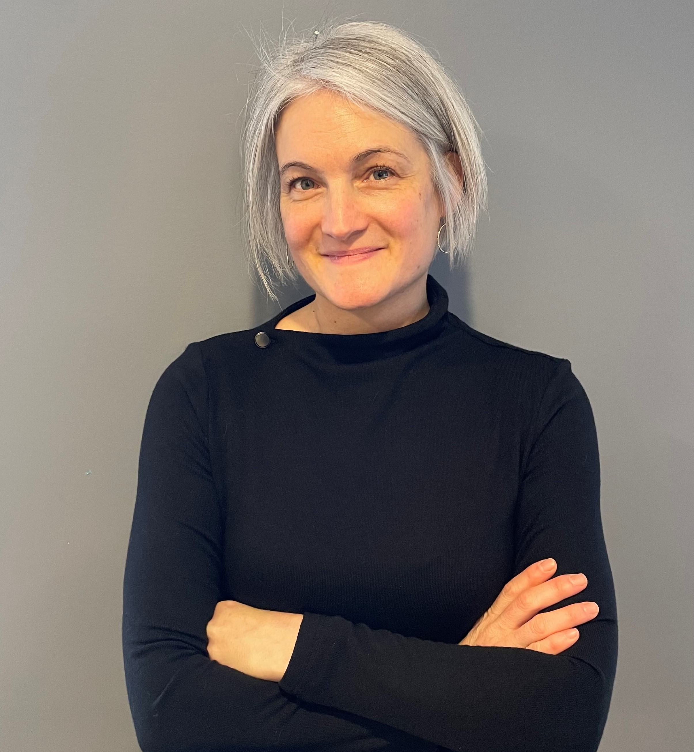 Headshot of Sarah Anderson, urban planner. Sarah is wearing a black shirt and is standing against a grey background with her arms crossed.