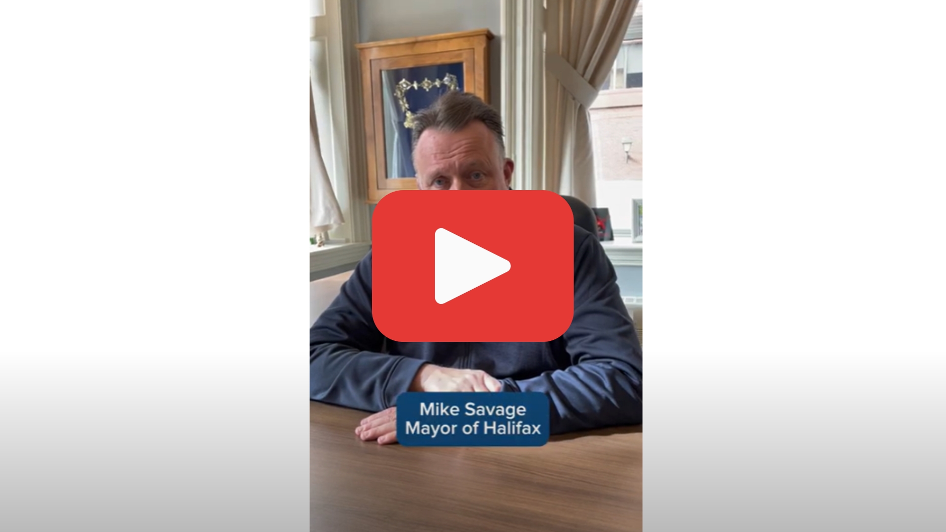 Video screenshot of Mayor Mike Savage sitting at his desk with a YouTube "play button" superimposed on him.