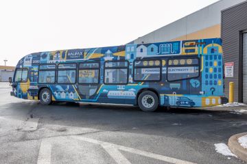 A photo of Halifax Transit’s pilot electric bus parked outside a bus garage. The bus is white on the front, like other Halifax Transit buses, but is wrapped in a special design on the back and sides. The design includes drawings of lighthouses, buses, ferries, the Citadel Hill clock tower, the Wave statue, seagulls, whales, lighting bolts, and more all connected by lines hinting at electric cables.