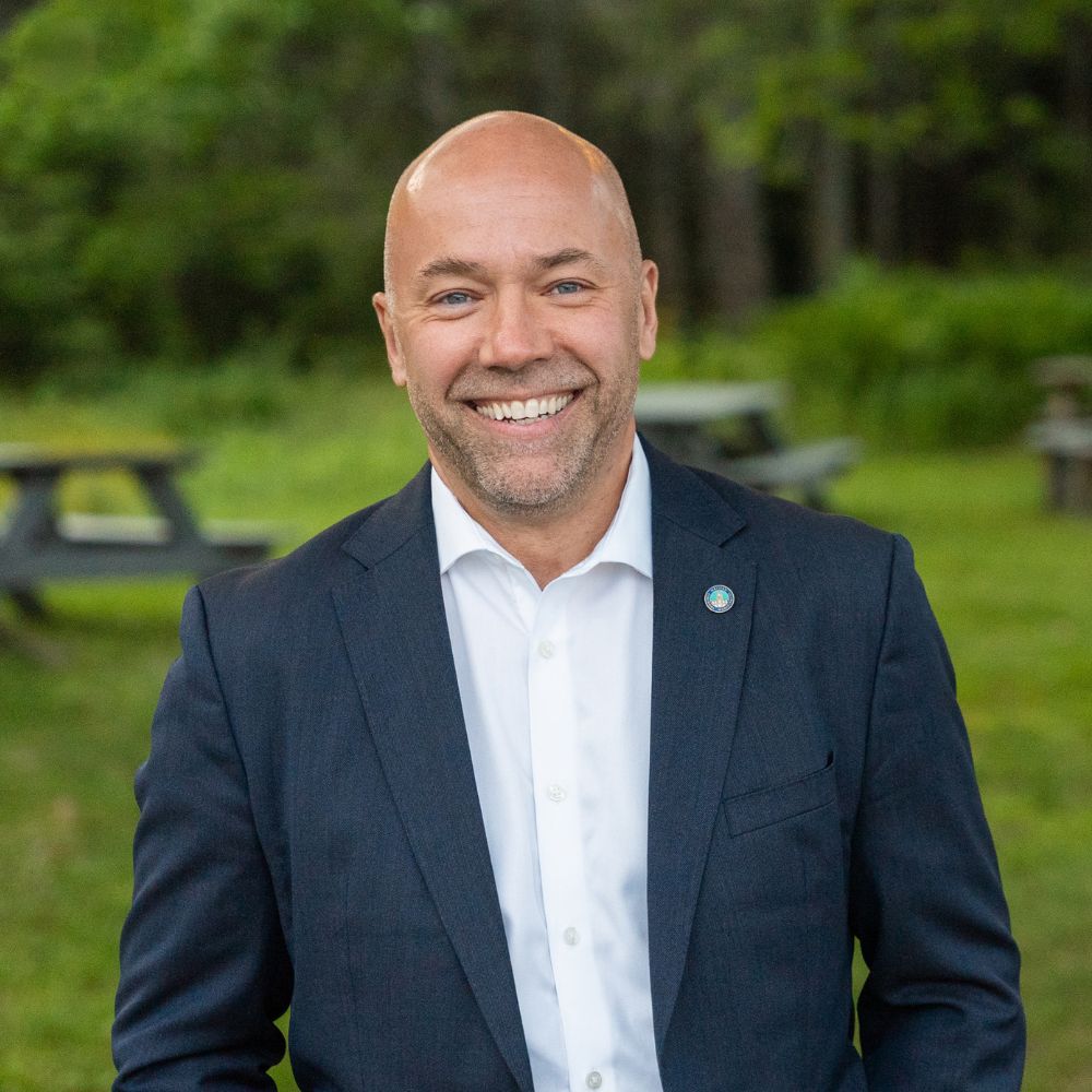 A man wearing a button-up shirt and dark sport jacket smiling at the camera.