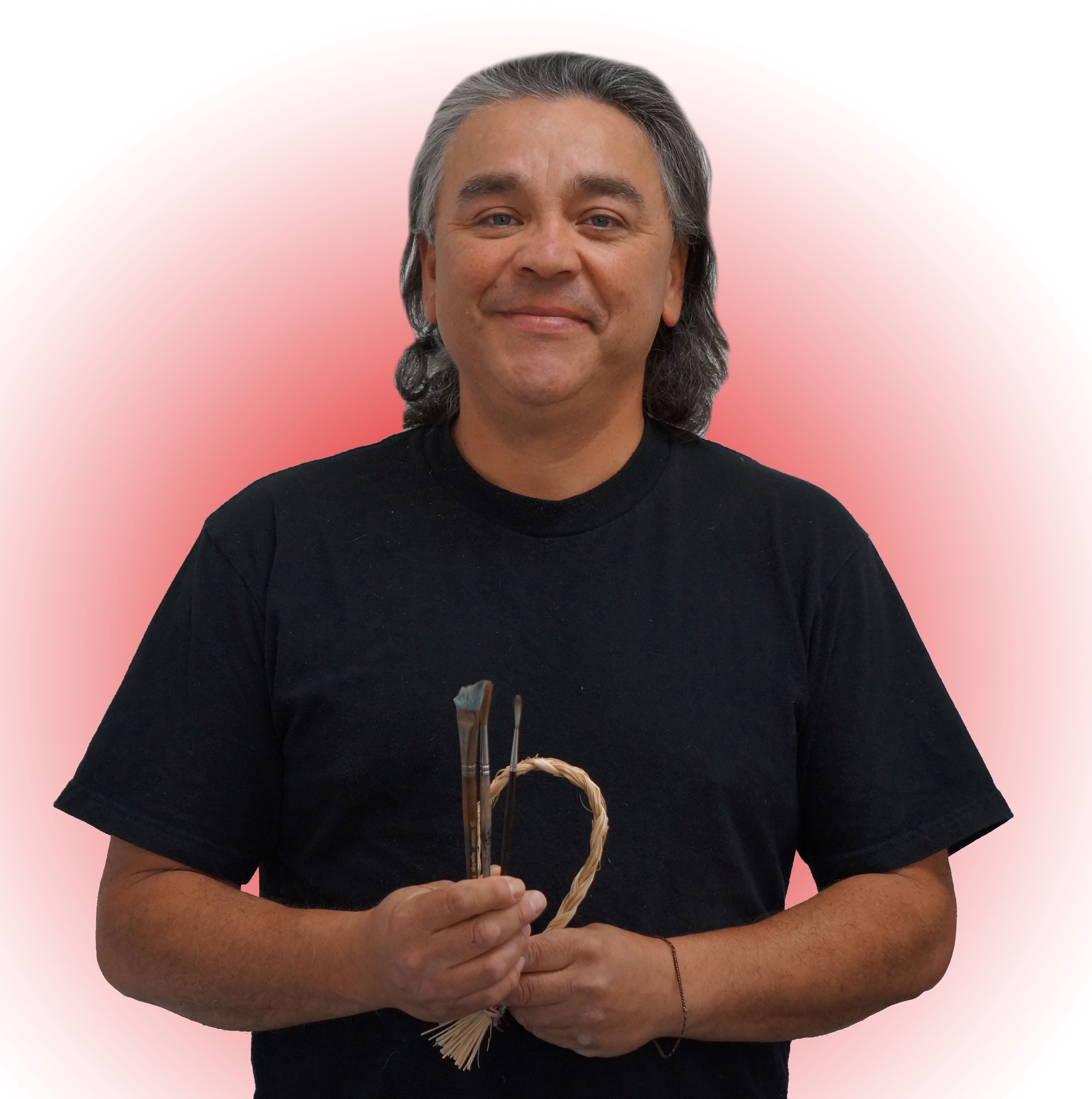 A photograph of Lorne Julien, a man in a black shirt holding paint brushes and sage.