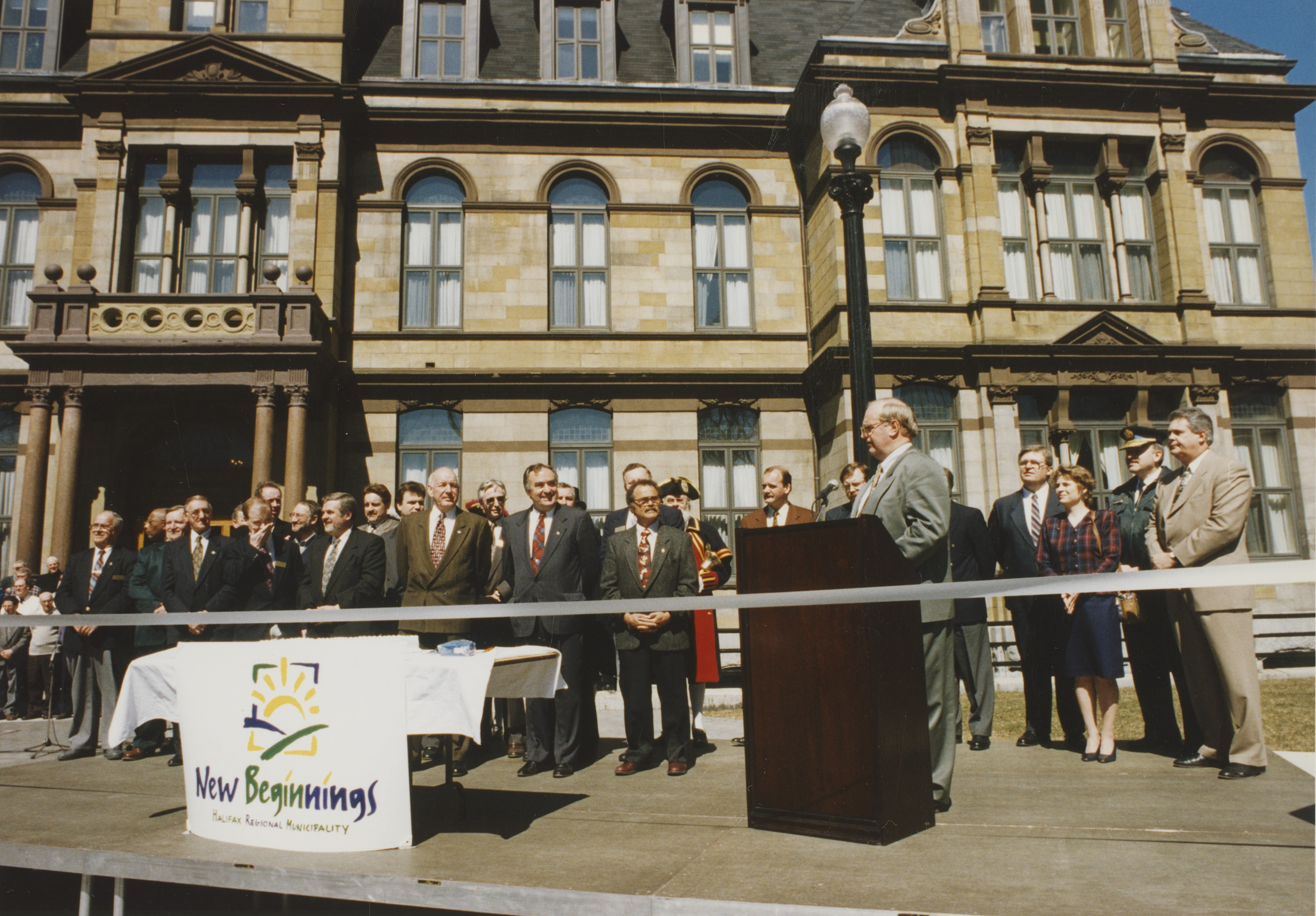 colour photo of crowd of officials gathered in front of City Hall with mayor Fitzgerald speaking at podium with a table labelled "New Beginnings" behind a ribbon