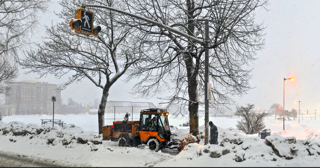 A snow plow working on sidewalk. Snow is falling and piling up high.