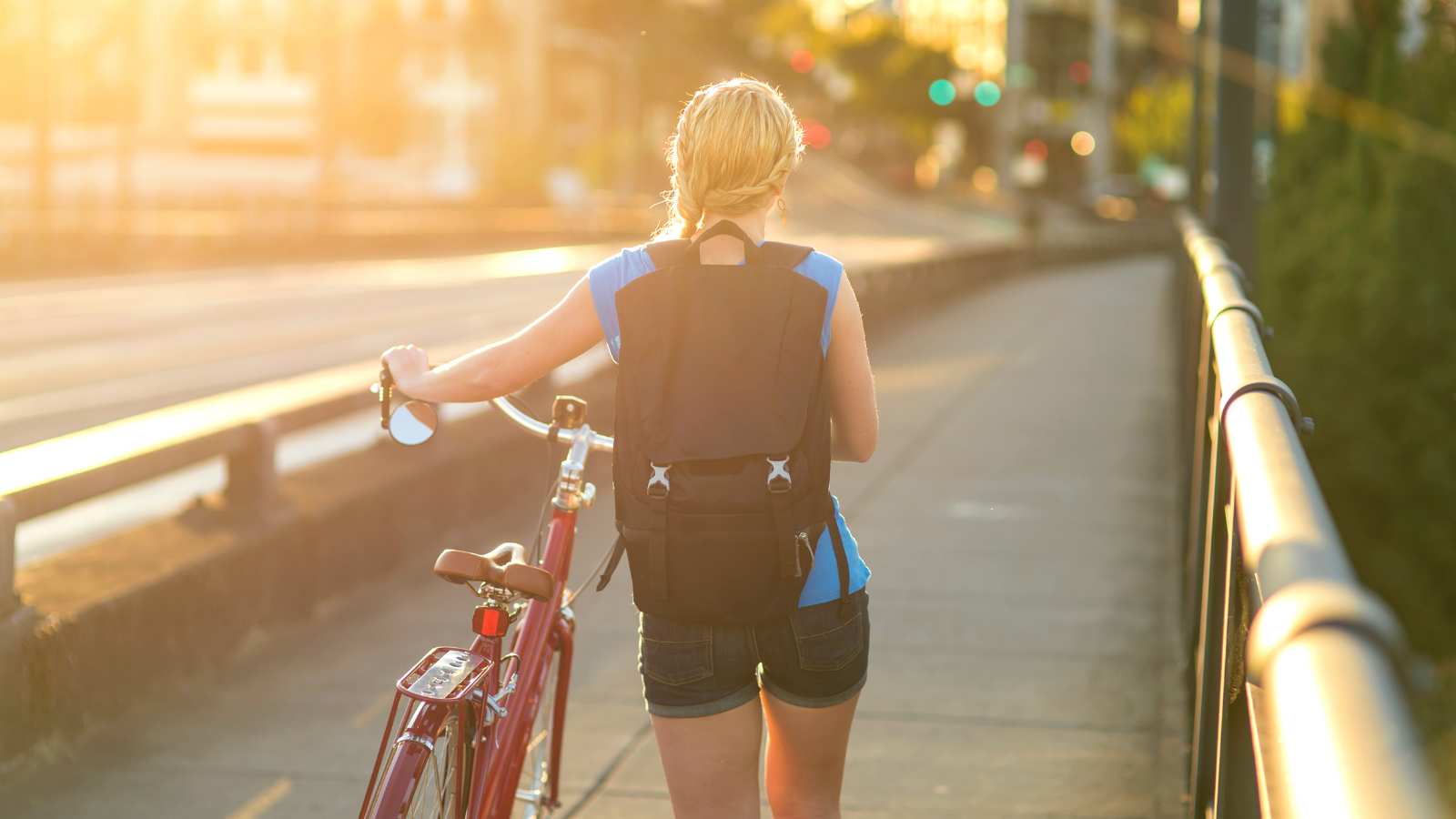A person with blond hair in a braid, wearing a backpack and shorts, walks a red bicycle across a bridge in the warm glow of the setting sun.