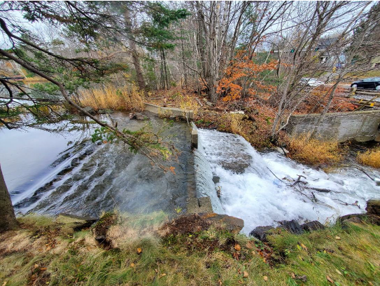 ID: Photo showing the Chocolate Lake Dam from the side, with the lake on the left and Herring Cove Road on the right. Water is flowing out of the lake toward the Northwest Arm.
