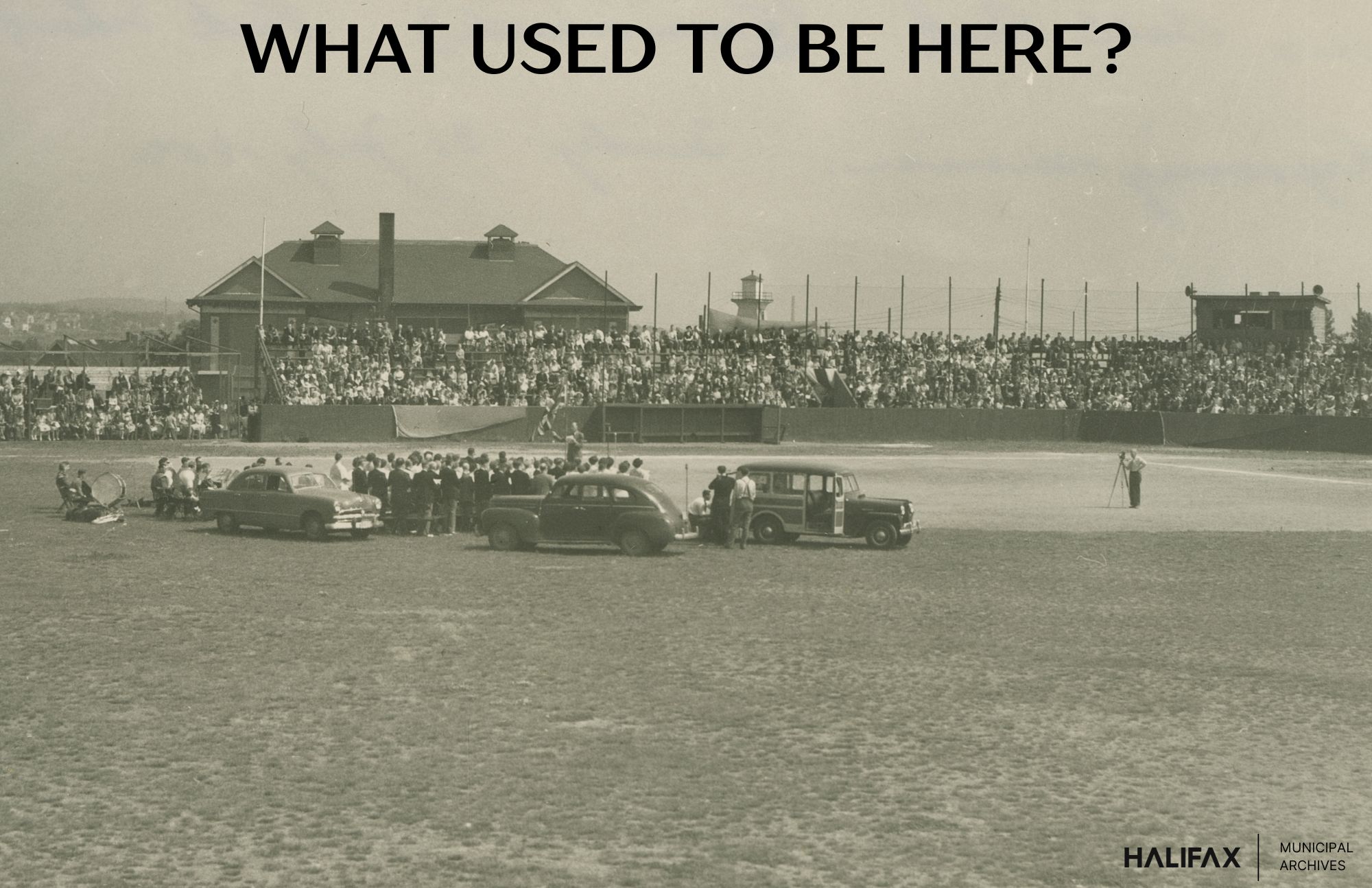 Black and white photograph of cars and people gathered in the middle of a baseball field with the crowd on bleachers watching.