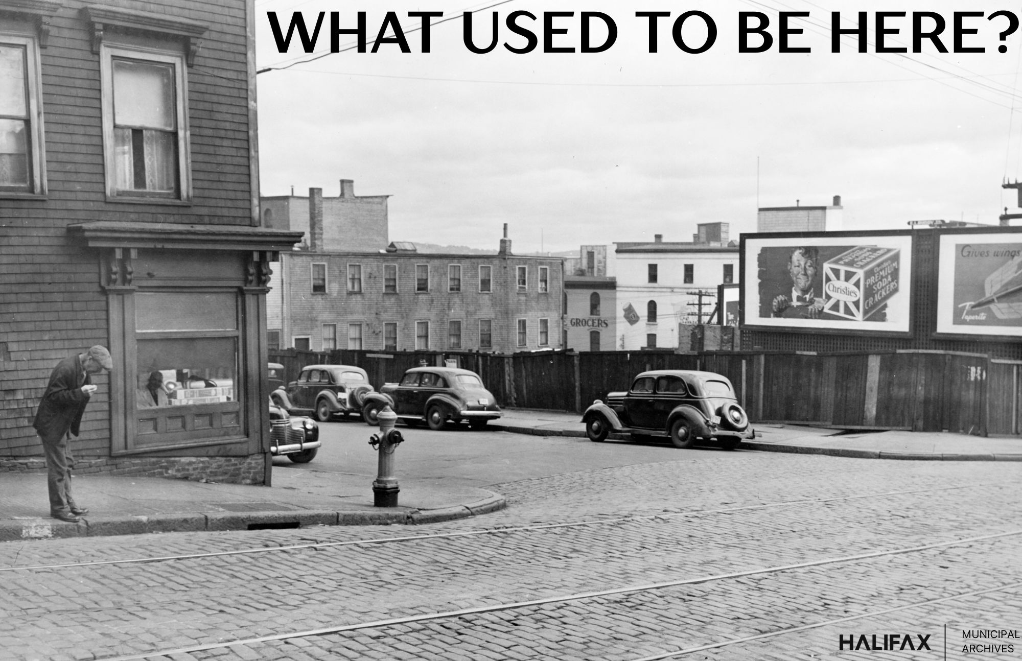 Black and white photo of a paving-stone street  with a store on the corner, cars parked on the street.