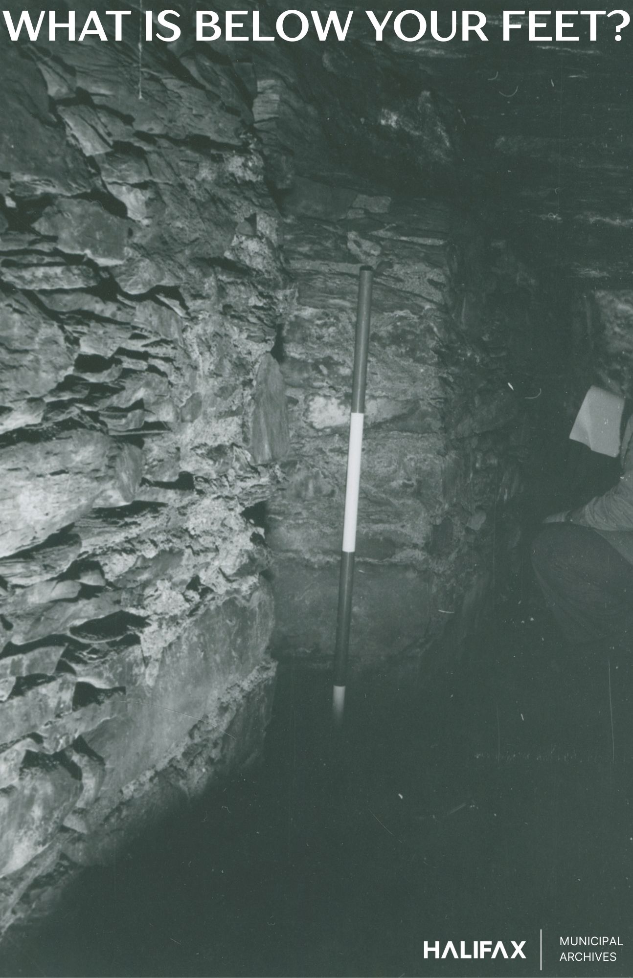 Dimly lit underground stone tunnel with rough, uneven walls and a damp floor. A vertical measuring stick is placed against the wall for scale. A person on the right holds a clipboard, suggesting documentation or research. Text reads “WHAT IS BELOW YOUR FEET?” at the top and “HALIFAX MUNICIPAL ARCHIVES” at the bottom right.