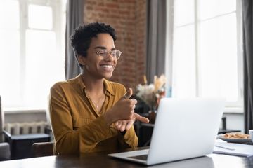 A person sits at a table using American Sign Language during a video call on a laptop.