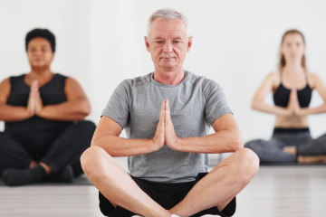 Older man and two other women sitting cross legged on the floor with hands in prayer pose