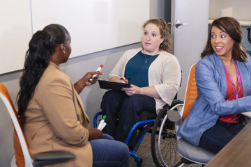 three people in a work meeting in front of a white board. The woman in the centre is a wheelchair user, and the other two women are sitting in office chairs. 