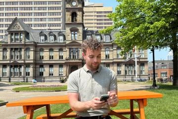 man sitting on an orange park bench in Grand Parade downtown Halifax glancing down at his phone; City Hall is in the background