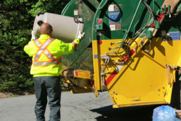 A worker wearing a high-vis vest tips a garbage bin into the back of a waste disposal truck.