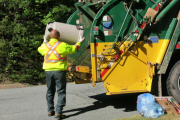 A worker wearing a high-vis vest tips a garbage bin into the back of a waste disposal truck.