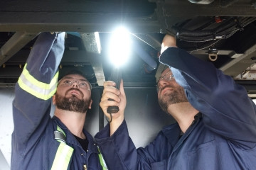 Two Transit mechanics looking up underneath a bus and doing some work. They are holding lights. 