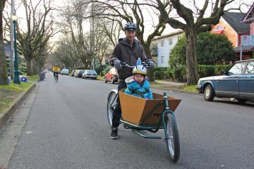 A father and son travel by cargo bicycle down a local street bikeway