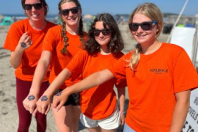 Four summer Rec staff members in matching orange work shirts with a local beach in the background.
